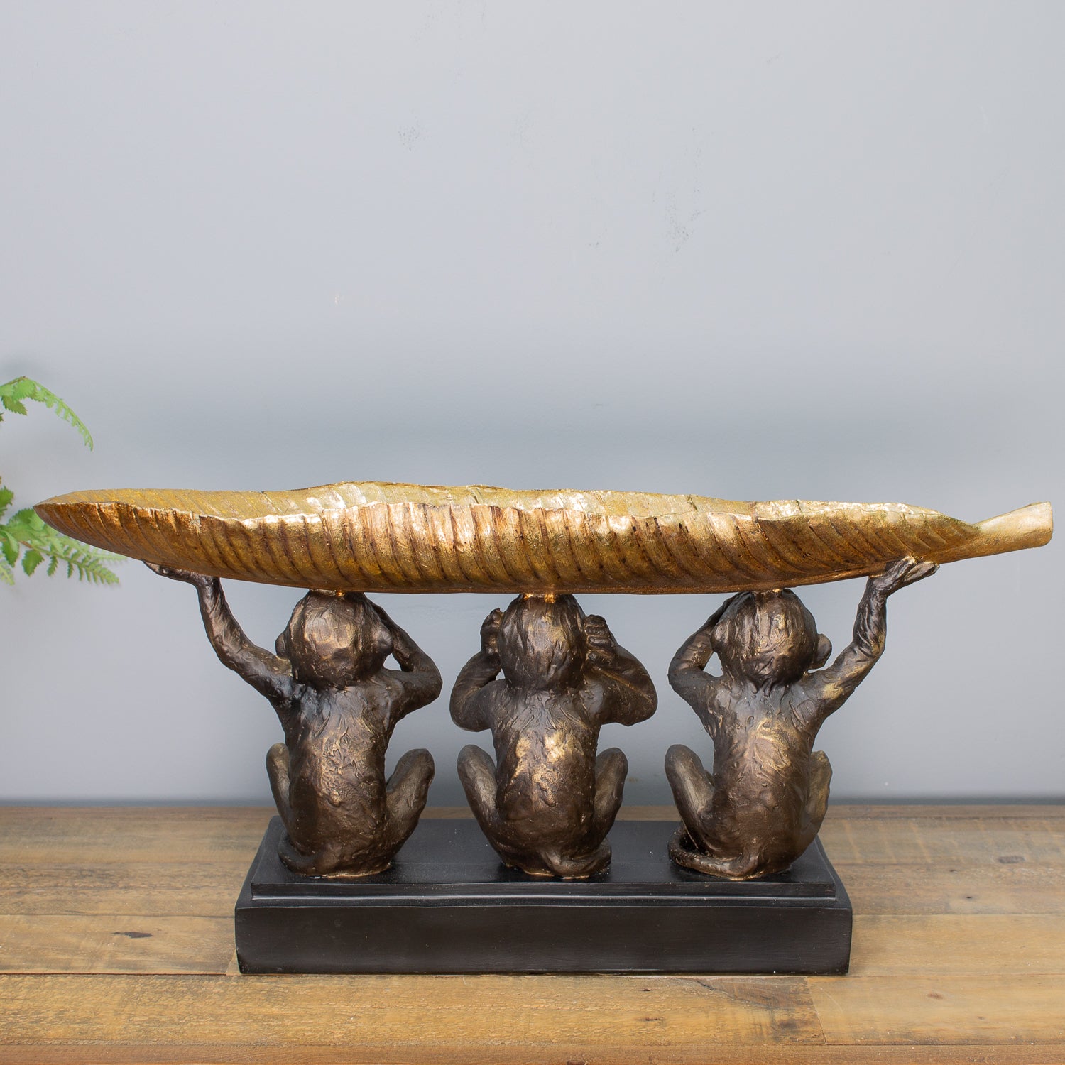 Bronze sculpture of three monkeys holding a large leaf on a wooden surface with a gray background